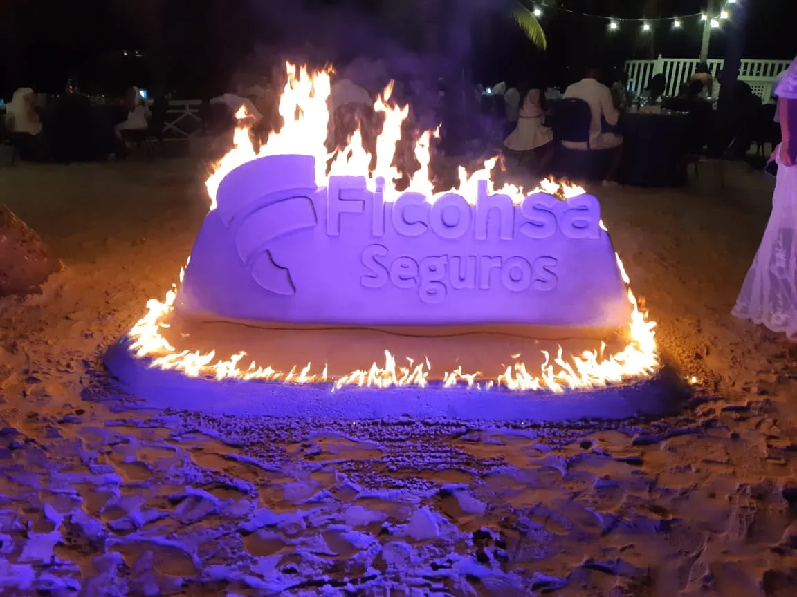 Sand sculpture of a company logo lit with fire at night, by Ronis Villeda in Roatan
